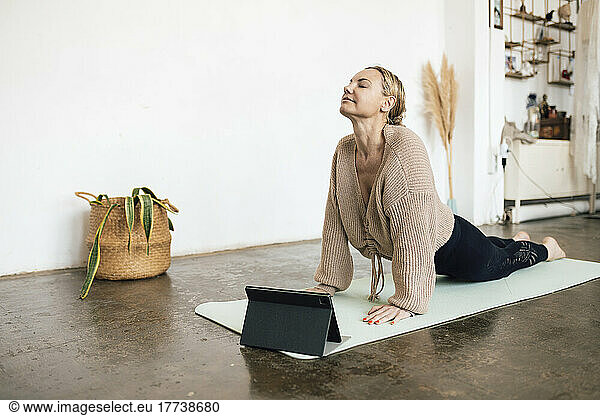 Woman practicing cobra pose on exercise mat in front of tablet PC at home