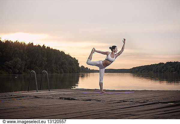 Woman practicing dance pose on pier by lake against cloudy sky during sunset