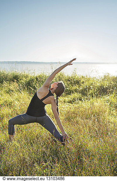 Woman practicing extended side angle pose yoga on grassy field by sea during sunny day