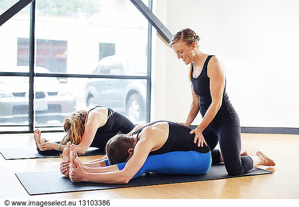 Woman practicing seated forward bend pose while instructor assisting friend at studio