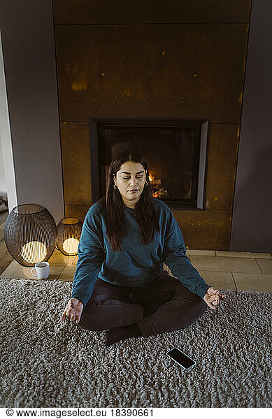 Woman practicing yoga while sitting cross-legged on carpet at home