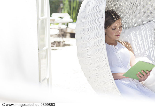 Woman reading book in hanging wicker chair outdoors