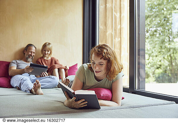 Woman reading book while father and daughter using digital tablet on bed at home