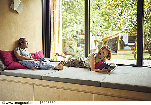 Woman reading book while man sitting with digital tablet on bed by window at home