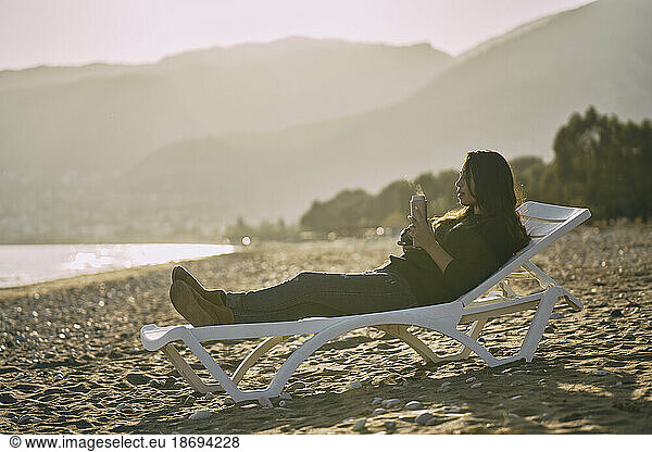 Woman reclining on deck chair holding thermos at beach