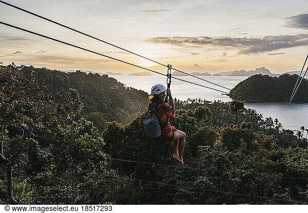 Woman riding on zip line over tree at sunset