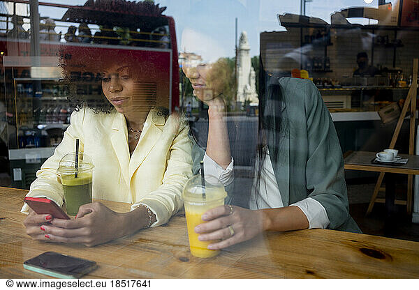 Woman sharing smart phone with friend sitting in cafe seen through window