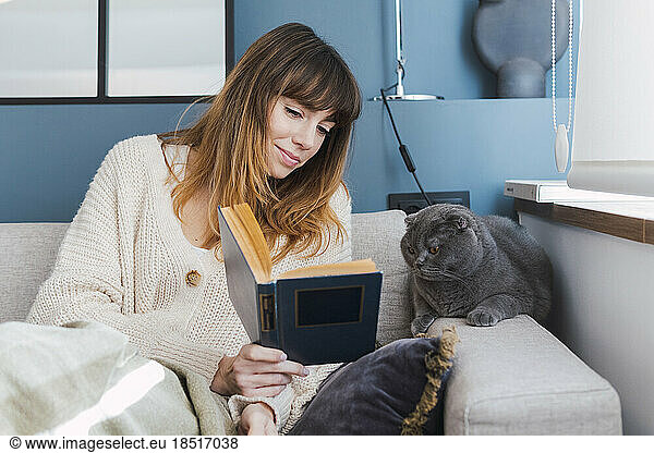 Woman showing book to cat sitting on sofa at home