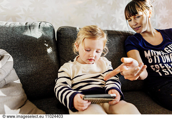 Woman showing girl to use smart phone while sitting on sofa