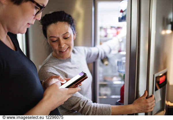 Woman showing mobile phone while girlfriend opening refrigerator in kitchen