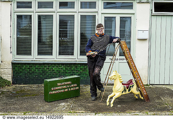 Woman sign writer and craftswoman holding paintbrush  palette and Maulstick  beside a ladder and small carousel horse.