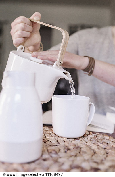 Woman sitting at a table in her apartment  pouring hot water into a mug  morning routine.