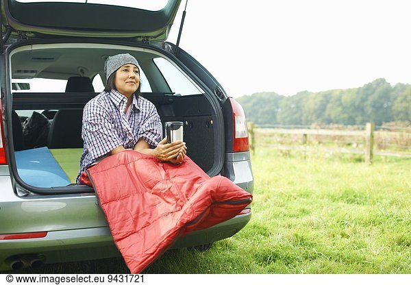 Woman sitting at rear of car with legs tucked in sleeping bag