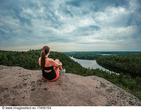 Woman sitting at summit of a hike overlooking lake and trees.