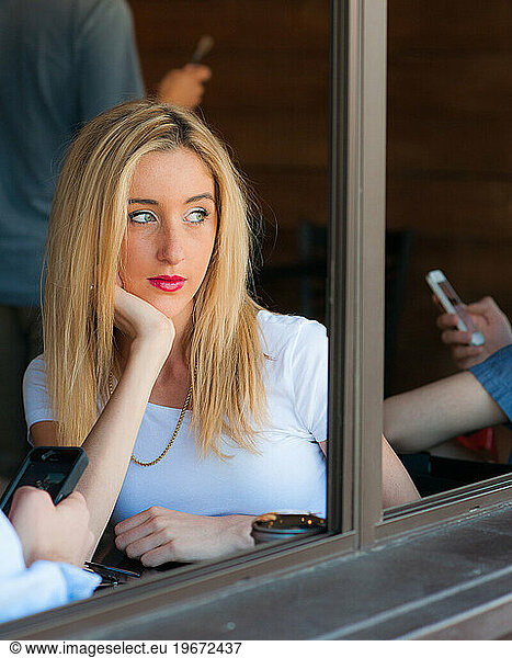 Woman sitting looking out of cafe window while other people use smartphones.