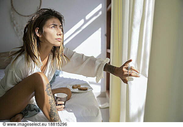 Woman sitting on bed and opening curtains at home