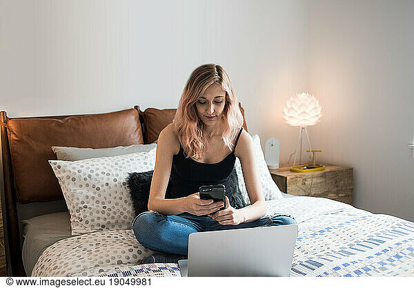Woman sitting on bed texting while using laptop