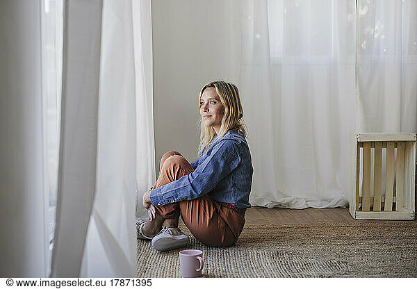 Woman sitting on carpet hugging knees near window at home