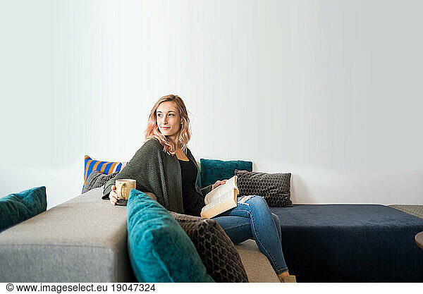 Woman sitting on couch with mug and book looking off camera