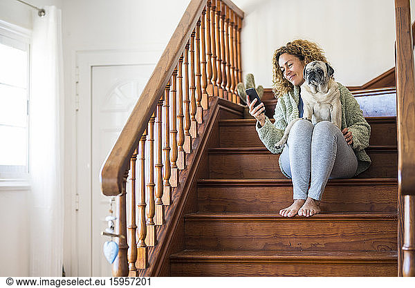 Woman sitting on stairs  using smartphone with pug on her lap