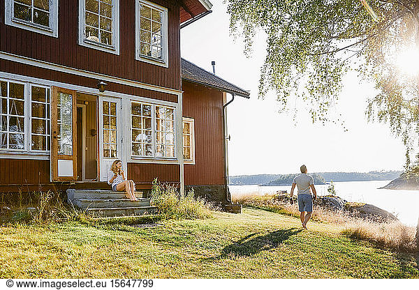 Woman sitting on steps at entrance of log cabin while man walking towards lake on sunny day