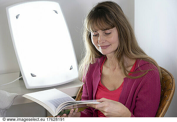 Woman sitting reading near a light therapy lamp.
