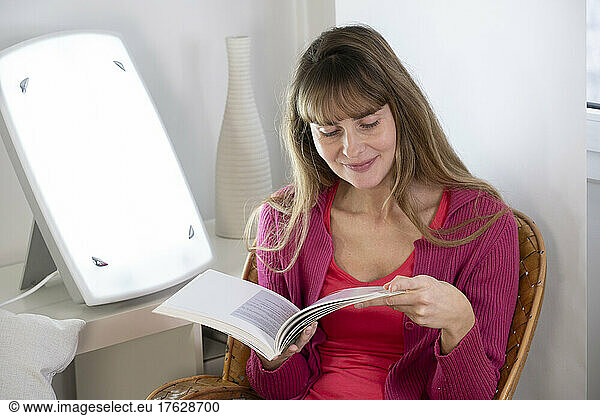 Woman sitting reading near a light therapy lamp.