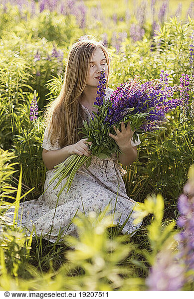 Woman smelling purple lupine flowers sitting in field