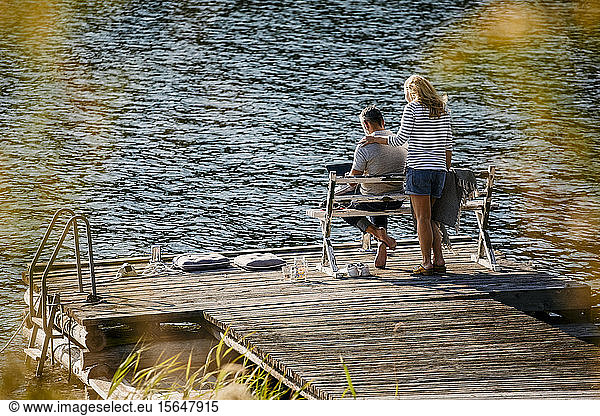 Woman standing by man working on laptop while female executive standing on wooden pier against lake