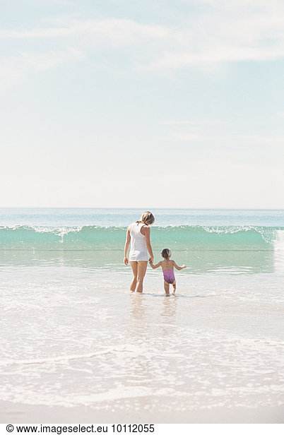 Woman standing hand in hand with her daughter on a sandy beach watching a wave.