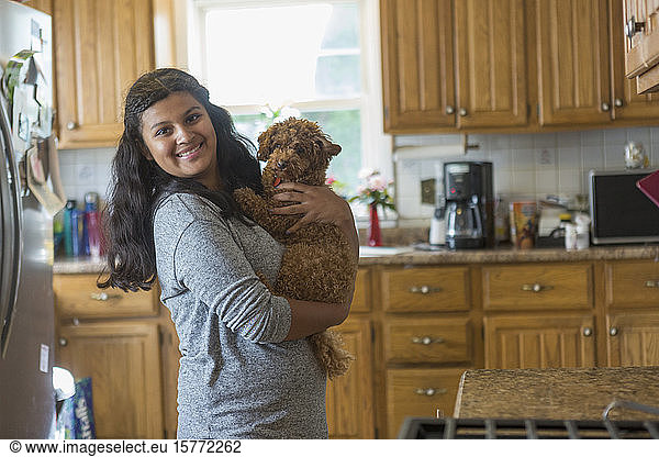Woman standing in the kitchen holding her pet dog and looking at the camera