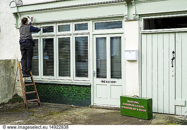 Woman standing on a ladder using paintbrush and maulstick  working on sign-writing architrave above a shop window.