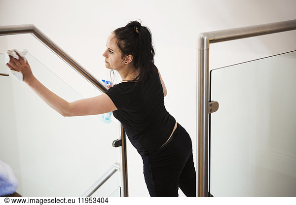 Woman standing on a staircase  cleaning a glass pane and banister rail.