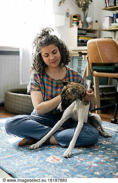 Woman stroking dog while sitting cross-legged on carpet at home