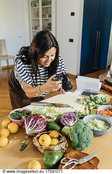 Woman taking picture of fresh vegetables using digital camera at home