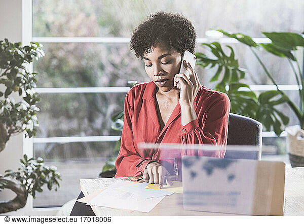 Woman talking on mobile phone and looking at documents at desk