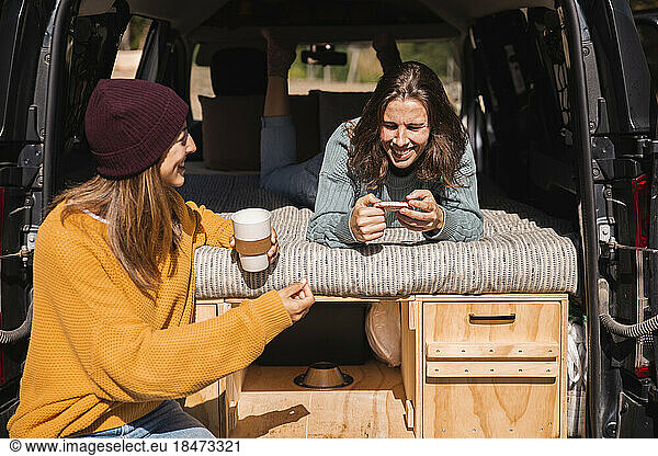Woman talking to smiling friend using smart phone lying in campervan