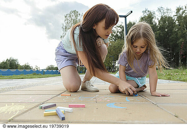 Woman teaching daughter to draw with chalk on tiled floor at park