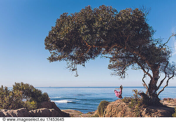 Woman tossing her dress under a tree on a cliff overlooking the ocean.
