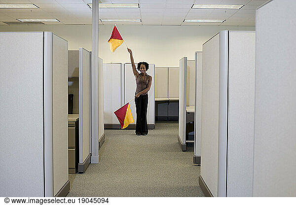 Woman uses semaphore flags to signal coworkers in cubicle-filled office.