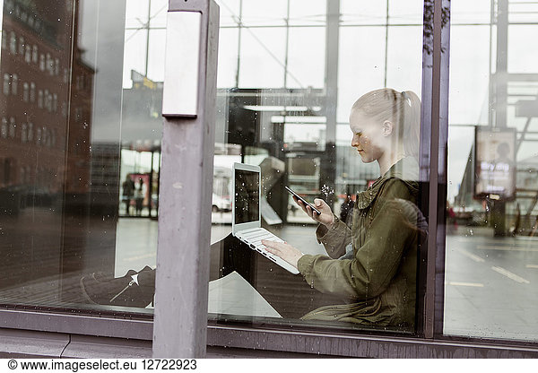 Woman using laptop and smart phone while sitting by window at station