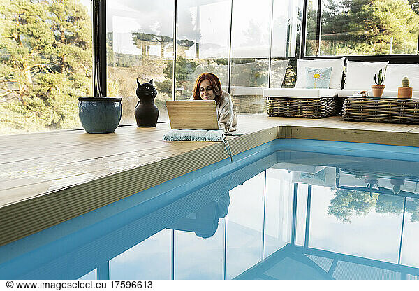 Woman using laptop by swimming pool in modern home