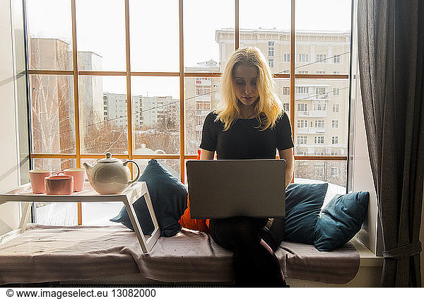 Woman using laptop computer by stool on alcove window seat at home