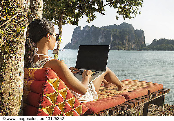 Woman using laptop computer while sitting on lounge chair against sea