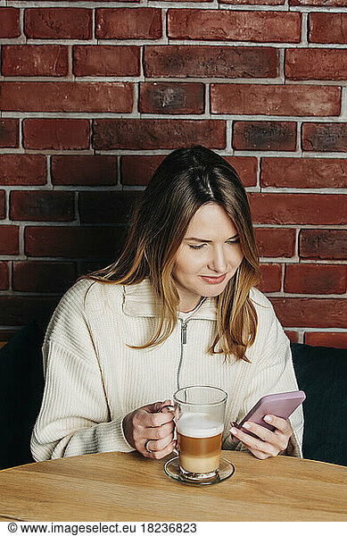 Woman using mobile phone and having cappuccino at table in cafe