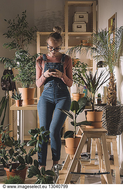Woman using mobile phone while leaning on table by potted plants at home