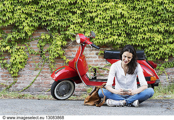 Woman using mobile phone while sitting against motor scooter and ivy wall