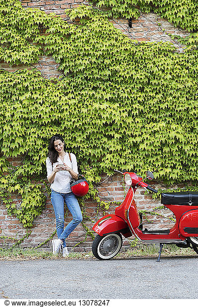 Woman using mobile phone while standing by motor scooter against ivy wall