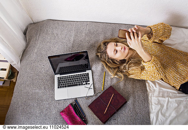Woman using phone while resting by laptop and book on bed