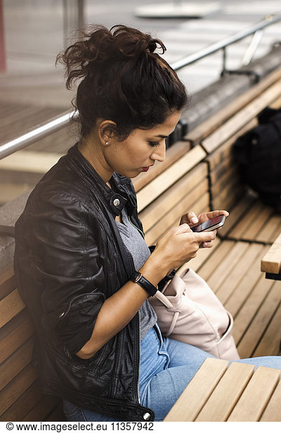 Woman using phone while sitting on wooden bench of sidewalk cafe in city